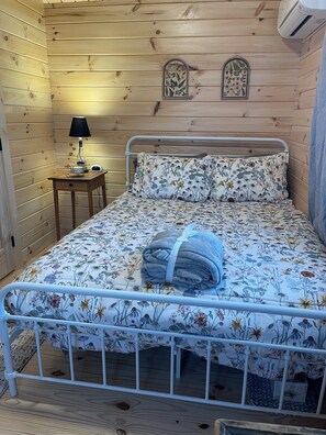 A wooden-paneled room with a metal bed frame, a bedside table with a lamp, and two wall-mounted air vents. - Cooperstown Double Play Glenview Cabin C (Cooperstown)
