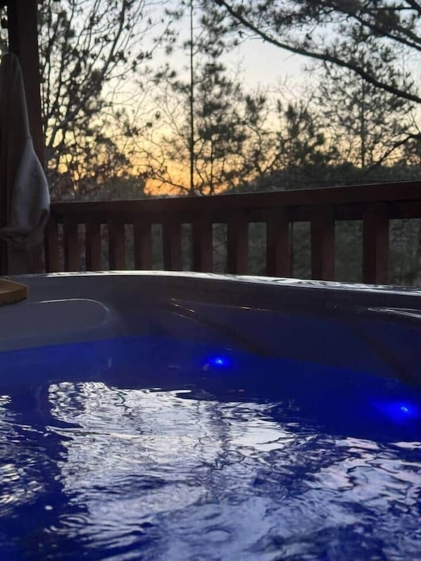 A hot tub with a wooden deck and a view of trees and a sunset.
