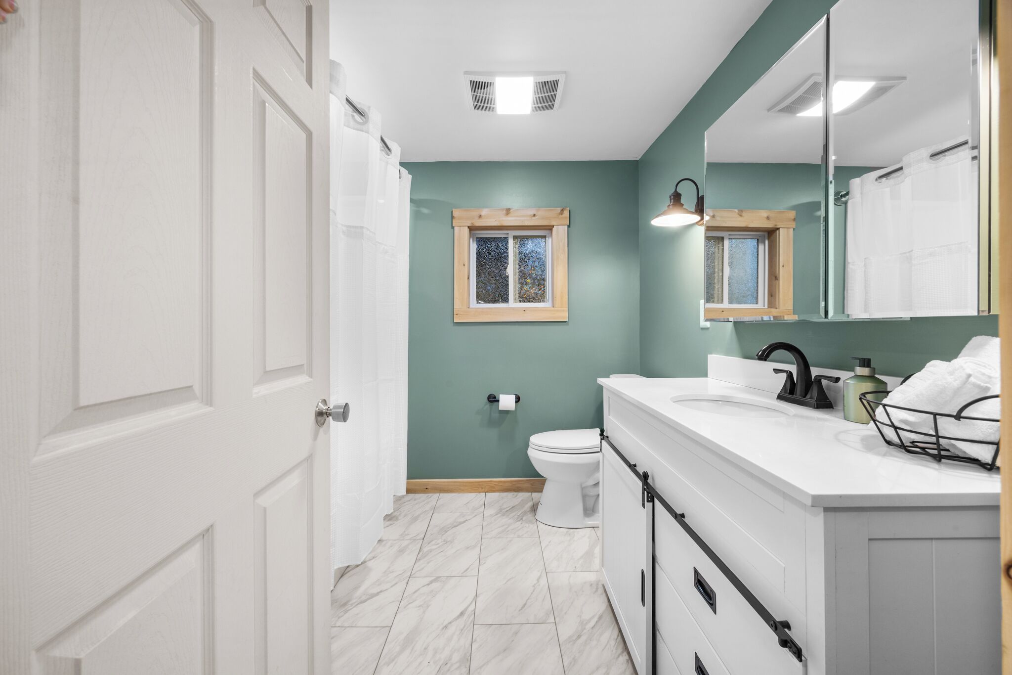 Une salle de bain avec des toilettes blanches, un lavabo avec un miroir et une fenêtre avec un cadre en bois.