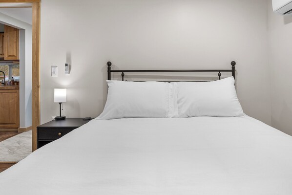 Una cama bien tendida con sábanas blancas, una mesita de noche con lámpara y un termostato fijado a la pared.