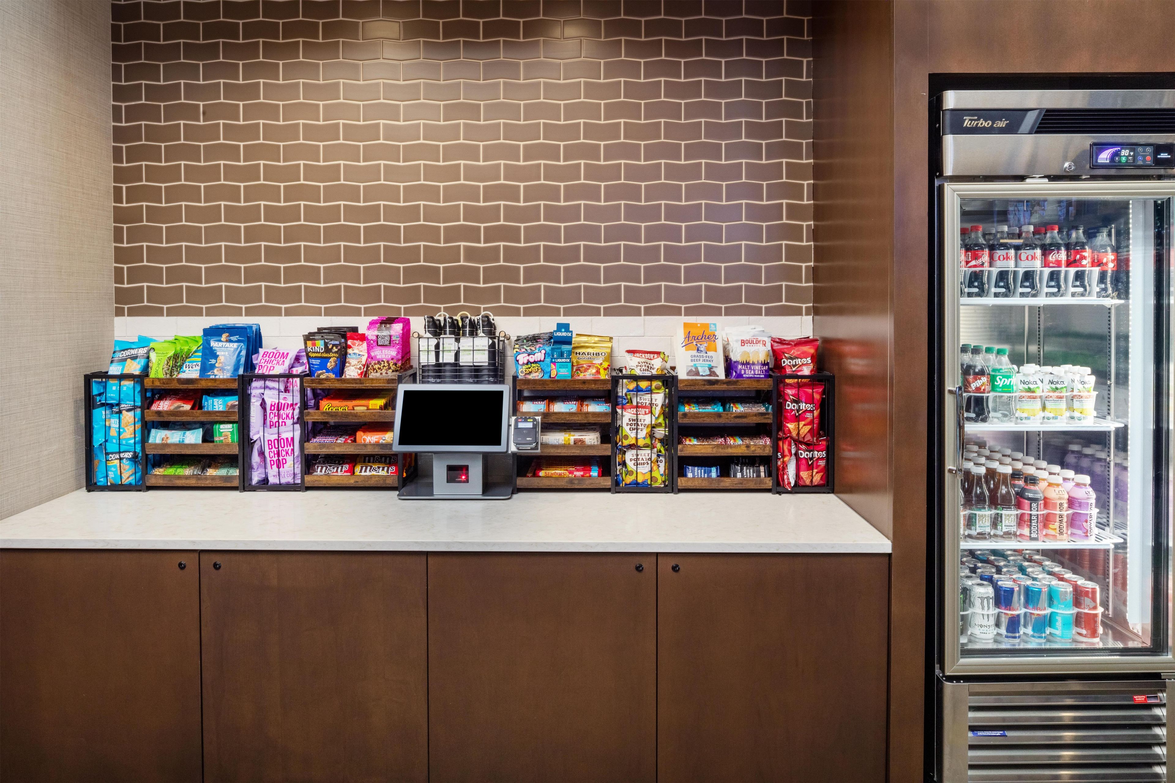 A self-service kiosk with a variety of snacks and beverages, including chips, cookies, and sodas, in front of a brick wall.