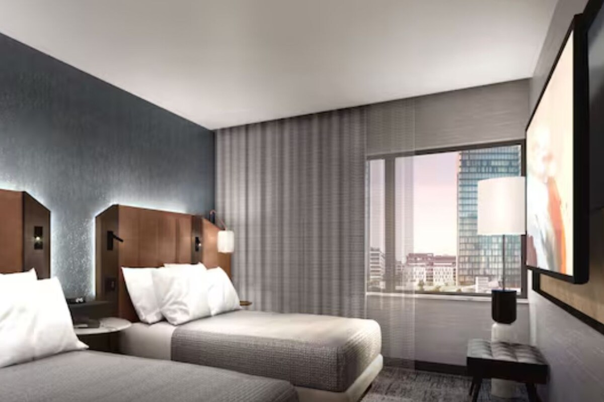 A hotel room with two beds, a large window with a city view, a flat-screen TV, and a modern design.