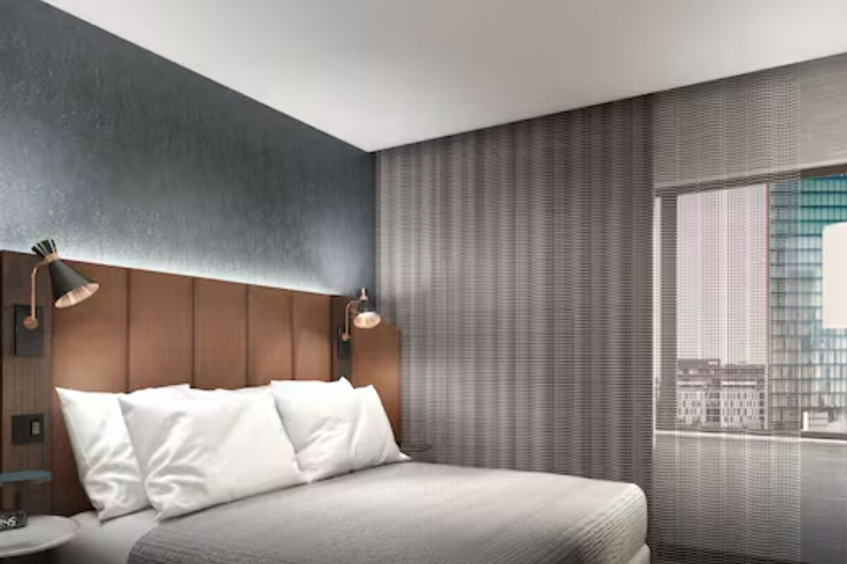 A modern hotel room with a bed, bedside lamps, a small table, and a large window with a city view.