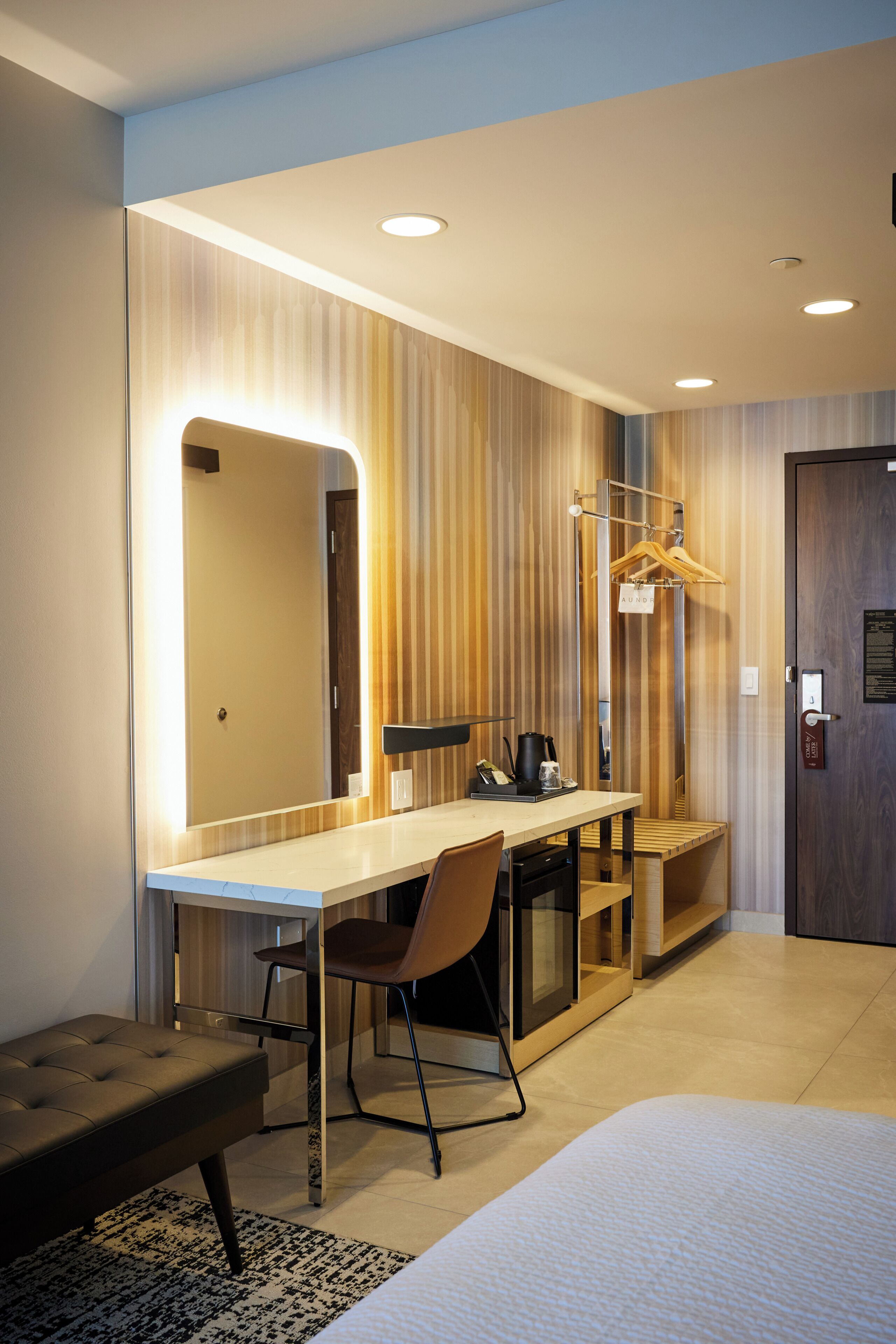A hotel room with a bed, a desk, a chair, a mirror, a hanging coat rack, and a door with a lock.