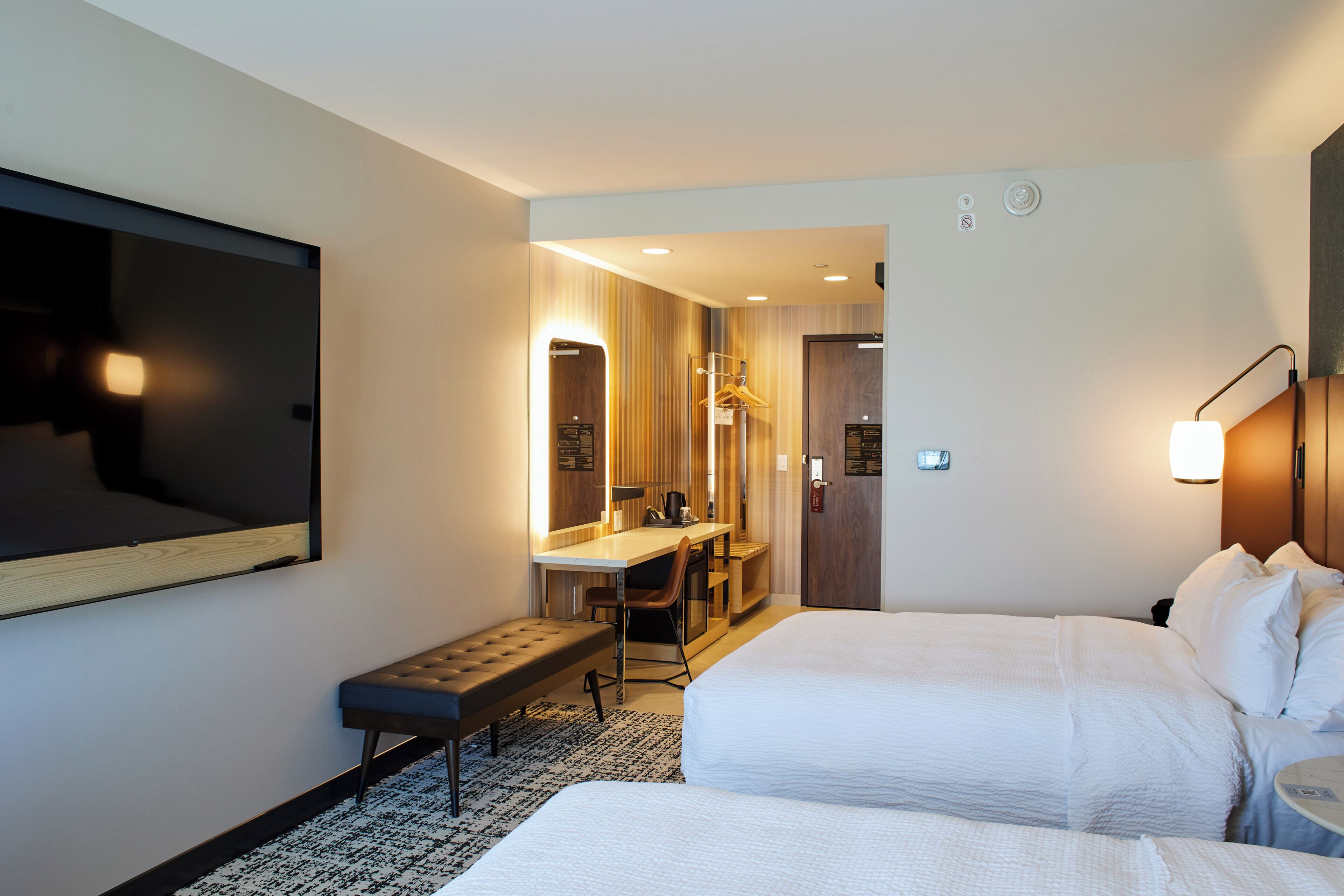 A hotel room with a bed, a desk with a chair, a bench, a TV, and a mirror.