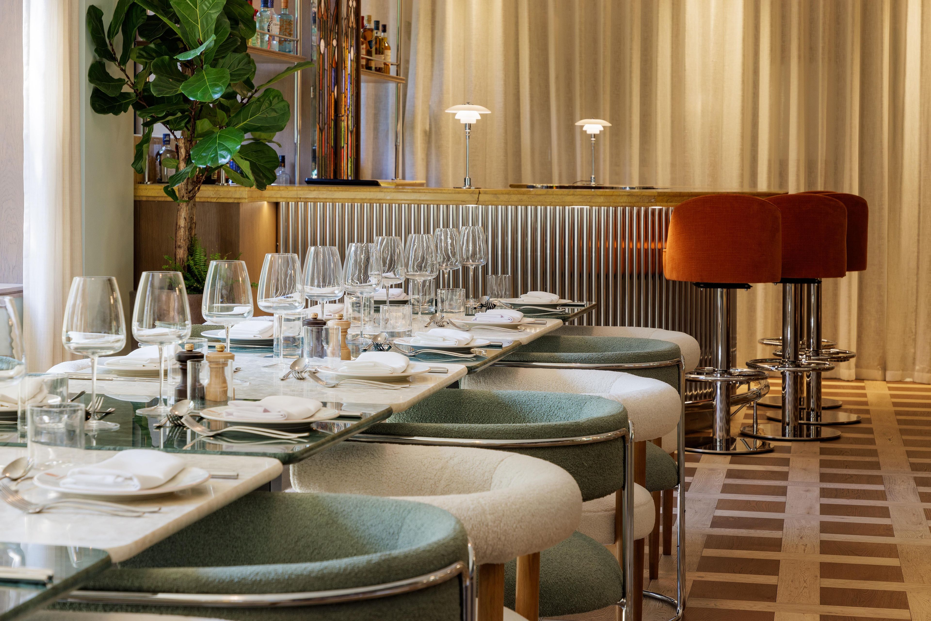 A restaurant with a long dining table set for multiple guests, a bar area with bottles, and a plant in the background.