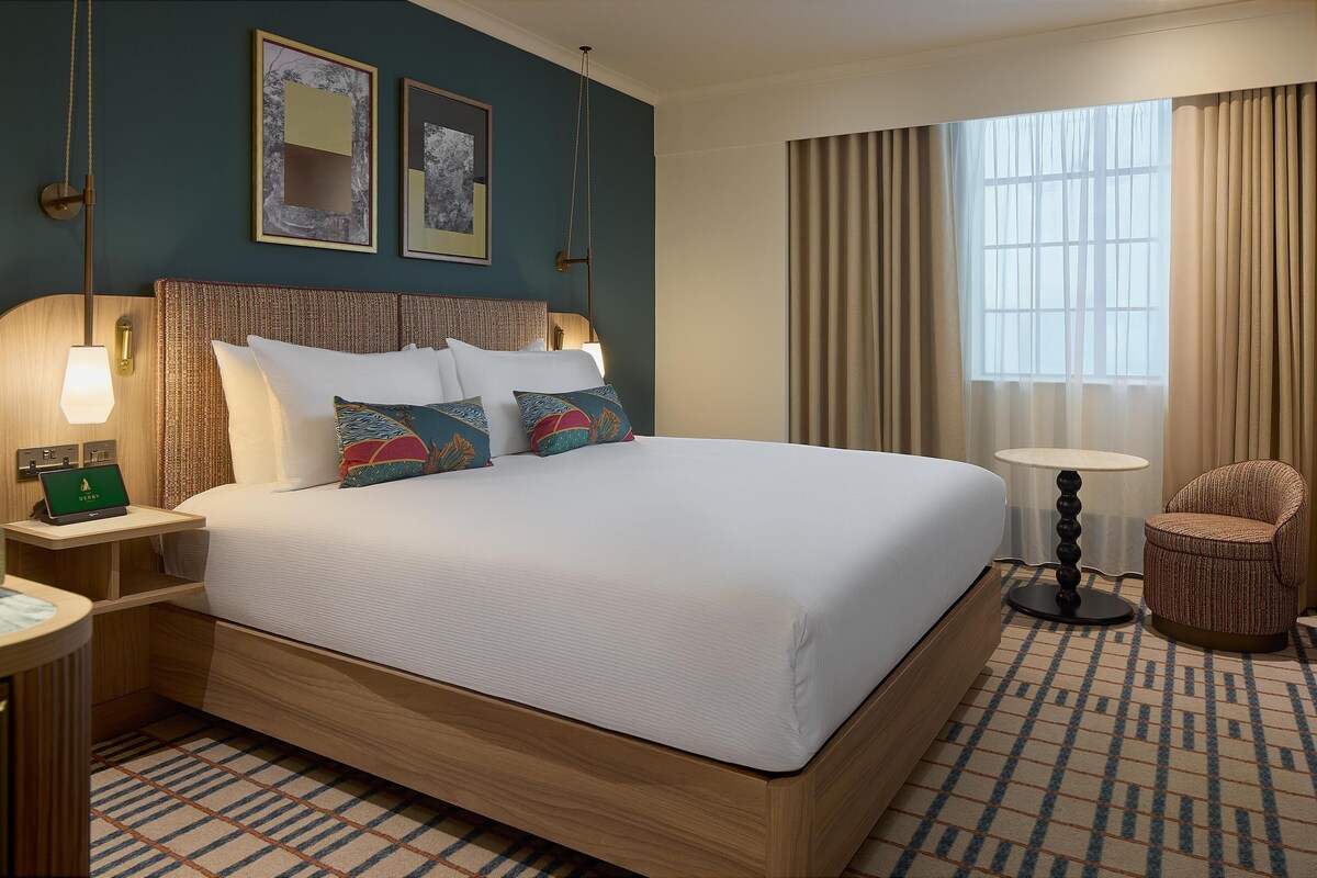 A hotel room with a large bed, a bedside table with a lamp, a chair, and a window with curtains.