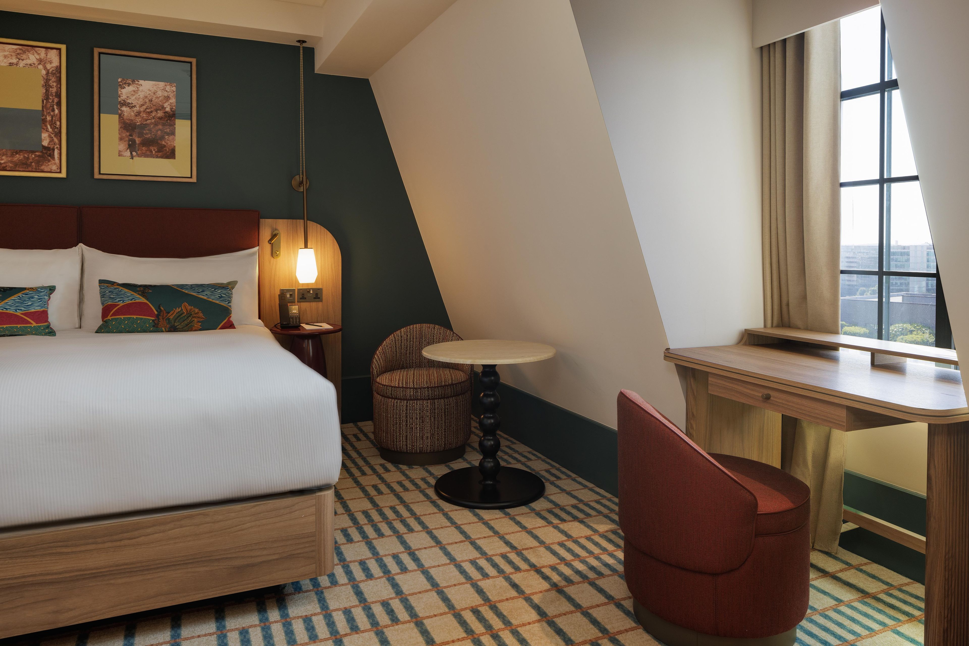 A hotel room with a bed, a desk, a chair, a small table, a lamp, and a window with a view.