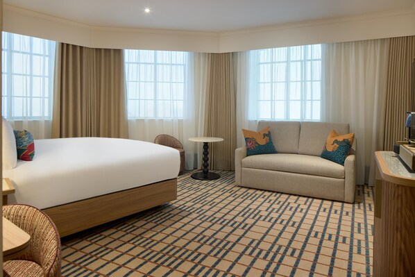 A hotel room with a large bed, a sofa, a small table, and a patterned carpet. - The Derby London City, Curio Collection By Hilton (London)
