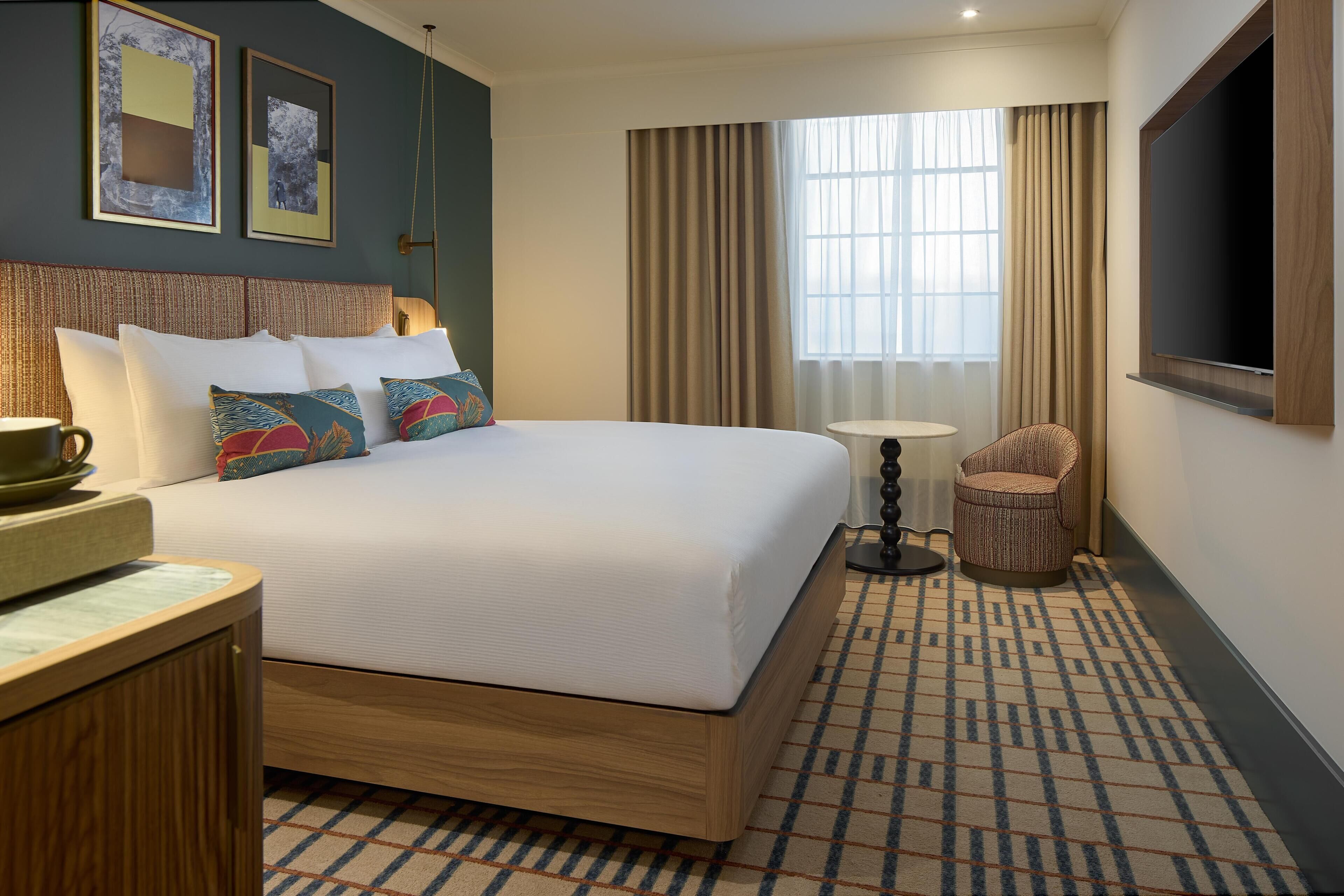 Una habitación de hotel con una cama grande, una mesita de noche, una silla, una pequeña mesa y un televisor montado en la pared.