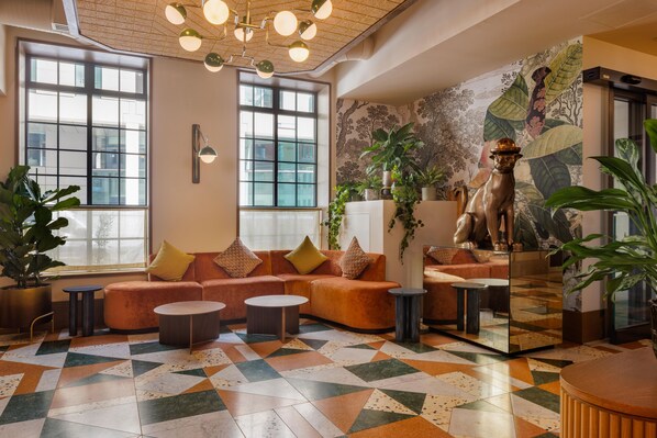 A modern lobby with a geometric patterned floor, a seating area with a sofa and armchairs, a decorative dog statue, and large windows allowing natural light.