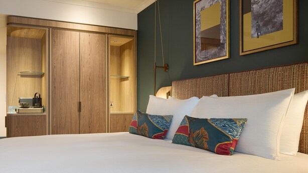 A hotel room with a large bed, wooden wardrobe, and a small bedside table with a lamp. - The Derby London City, Curio Collection By Hilton (London)