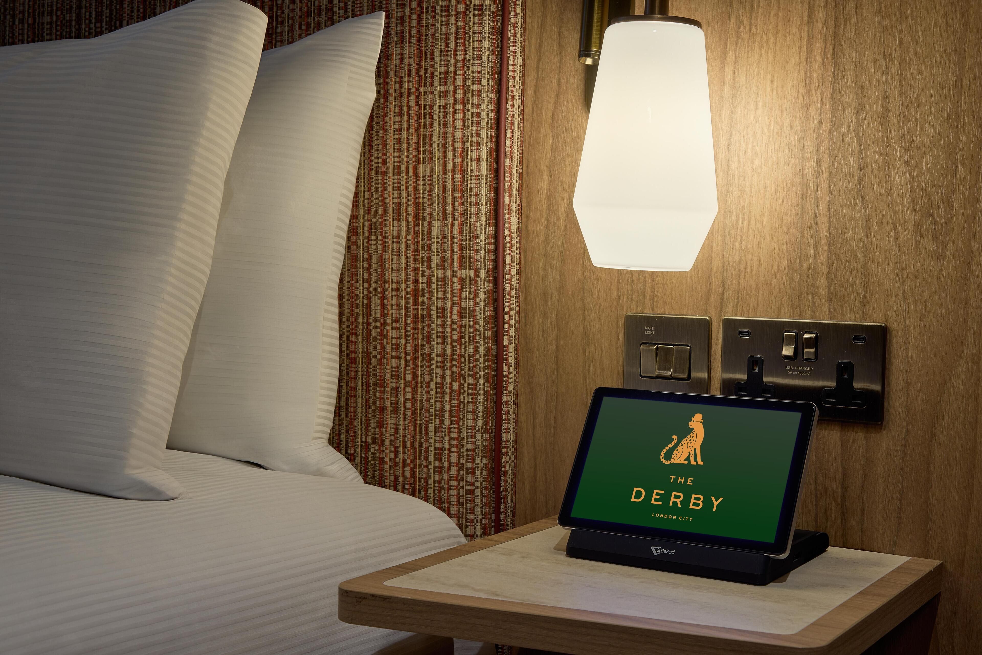 Quarto de hotel com cama bem arrumada, mesa de cabeceira com um laptop exibindo 'The Derby London City' e luminária fixada na parede.