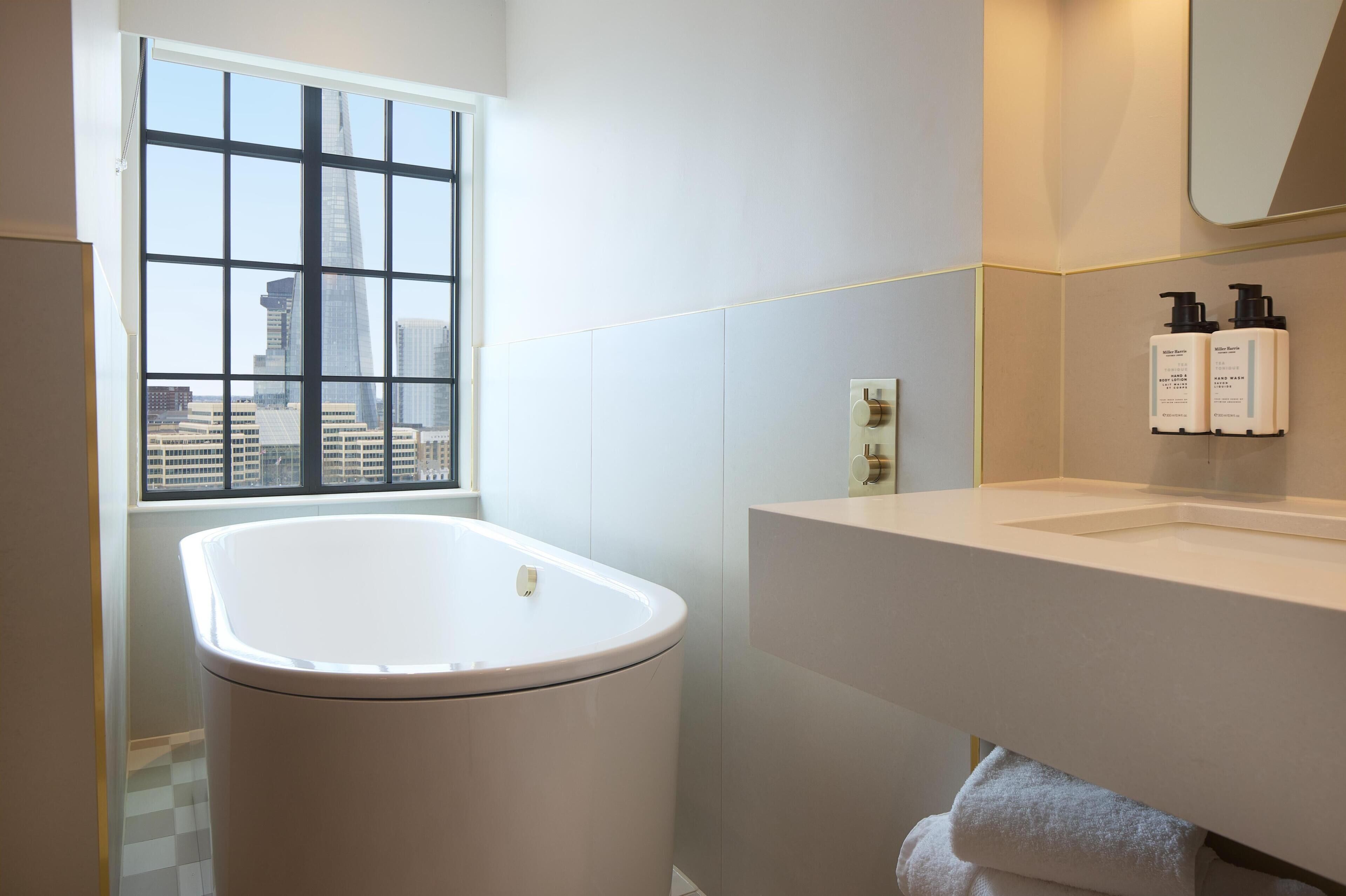 Un baño moderno con bañera, una ventana con vista a la ciudad, un lavabo y artículos de tocador en la pared.