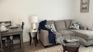 Un salon avec un canapé gris, une table basse en verre, une table à manger sur laquelle sont empilées des tasses blanches, une lampe et un tableau encadré accroché au mur.