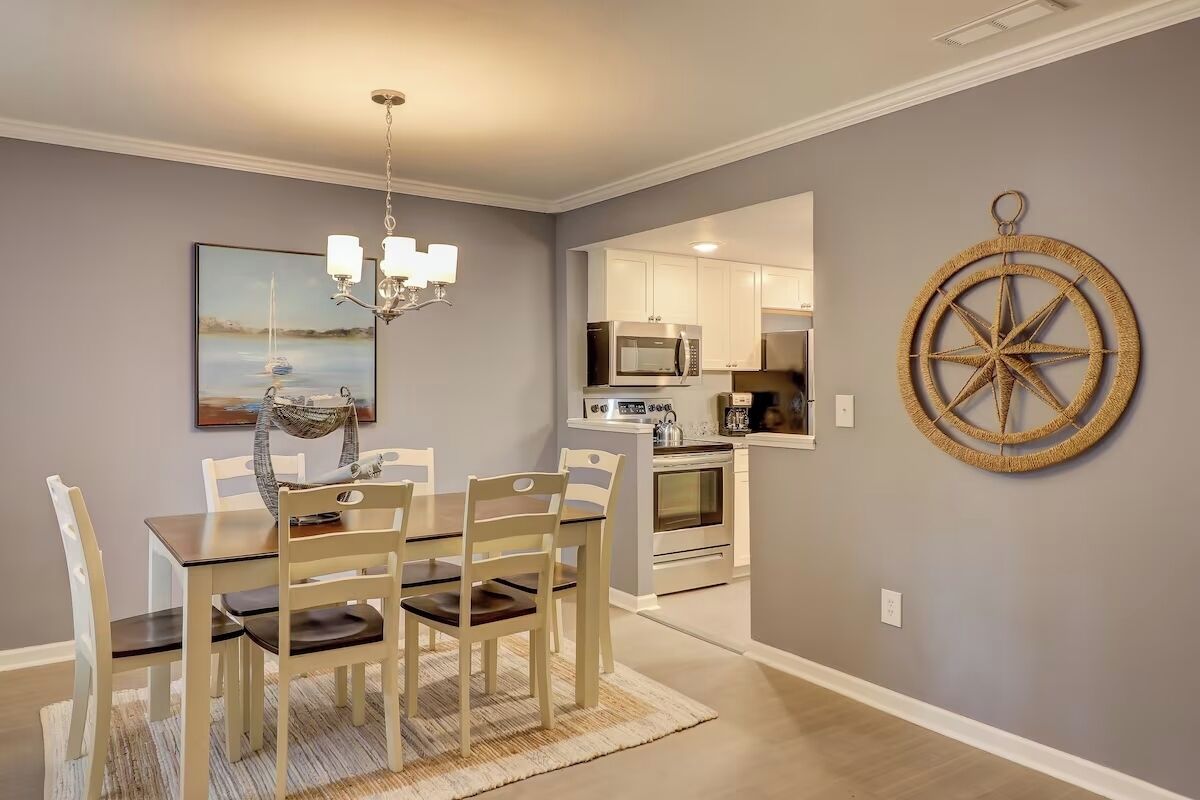 A dining area with a table and chairs, a chandelier, a painting, and a kitchen in the background.