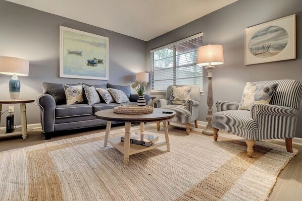 A living room with a grey sofa, striped armchair, and a round coffee table. There are two lamps, a painting, and a window with blinds. - Spacious 3BR Hilton Head Getaway + Pool Access (Hilton Head Island)