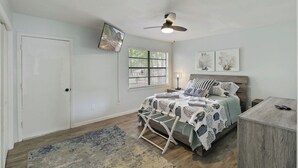 A bedroom with a bed, a ceiling fan, a window with blinds, a nightstand, and a desk.