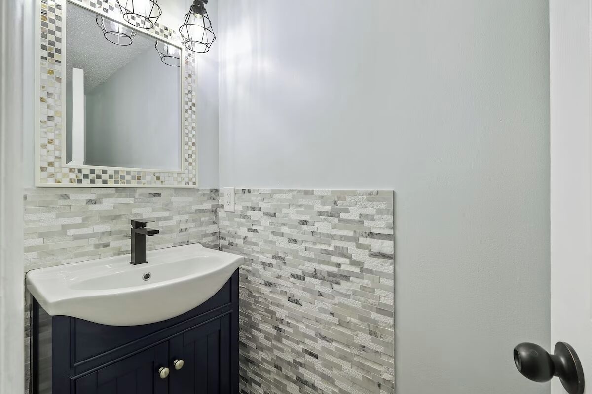Un baño con lavamanos blanco, muebles oscuros, salpicadero de azulejos y un espejo con marco geométrico.