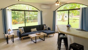 A modern living room with a sofa, coffee table, and bar area. There are large windows with curtains, a ceiling fan, and an air conditioning unit.