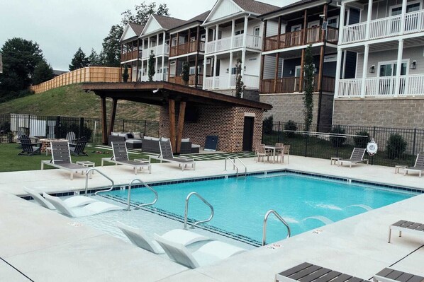 Un área de piscina con sillones de descanso, un espacio de estar cubierto y un edificio con balcones.