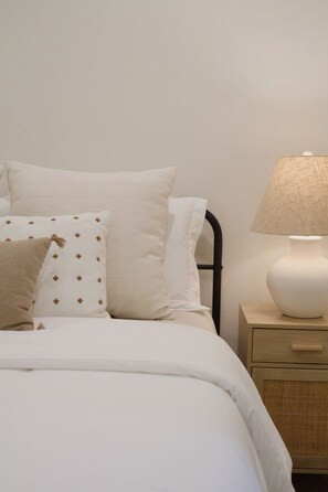 Una cama con ropa blanca y dos almohadas, una con estampado, una mesita de noche con lámpara y un mueble con cajones.