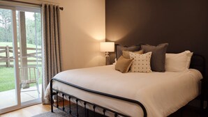 Un dormitorio con cama, almohadas, una mesita de noche, una lámpara y vistas a una terraza de madera y follaje exterior.