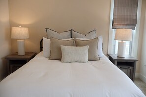 Una cama bien arreglada con varios almohadones, dos mesitas de noche con lámparas y una ventana con cortina.