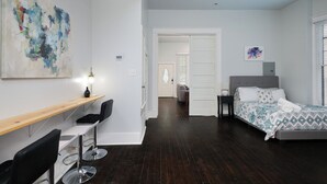Une chambre moderne avec un lit, un bureau et une chaise, un ventilateur de plafond, et un couloir menant à une autre pièce.