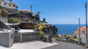 Zona residencial costera con entrada vigilada, un coche estacionado en el interior y vistas al mar.