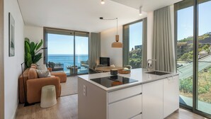 Una cocina moderna con una isla central, ventanas grandes con cortinas, un comedor con vistas al mar y una sala de estar con sofá y planta.