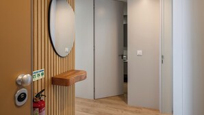 Habitación de hotel con pared de paneles de madera, un espejo redondo, estante de madera, extintor y puerta que da a otra habitación.