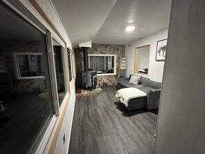 A living room with a stone wall, a grey sofa, a wooden floor, and a large window.