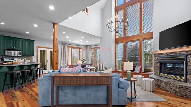 A spacious living area with a fireplace, a kitchen with green cabinets, and a large window. - Helen Haus: Golf Sim & Hot Tub Theater Retreat (Helen)