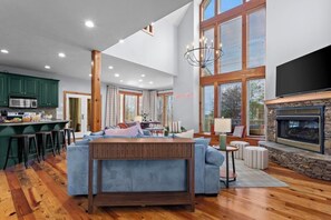 A spacious living area with a fireplace, a kitchen with green cabinets, and a large window. - Helen Haus: Golf Sim & Hot Tub Theater Retreat (Helen)