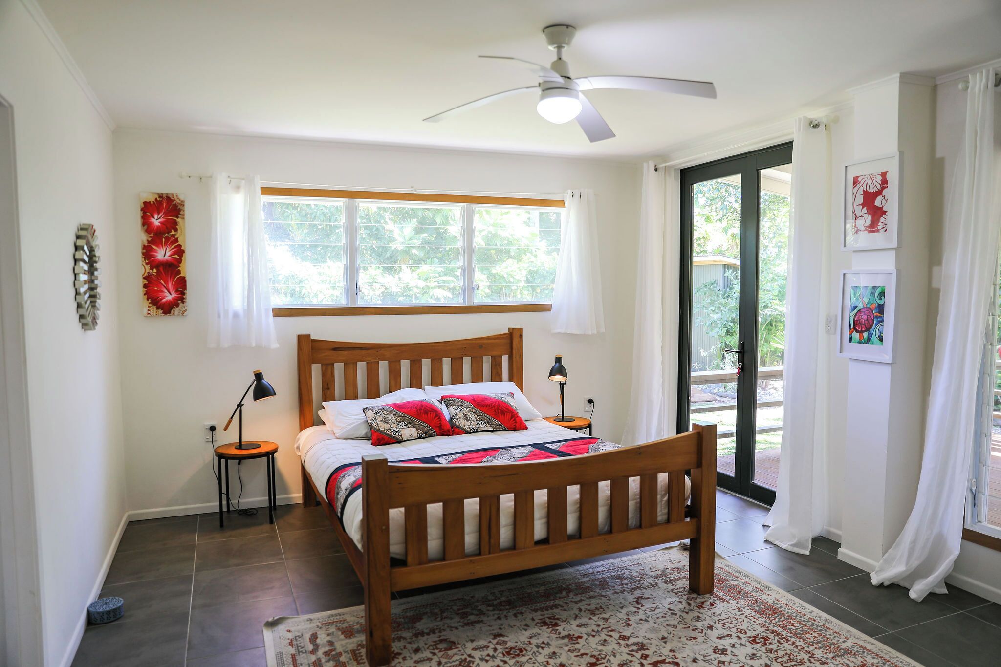Quarto com cama de madeira, ventoinha no teto, janela com cortinas, mesa pequena com abajur e tapete.