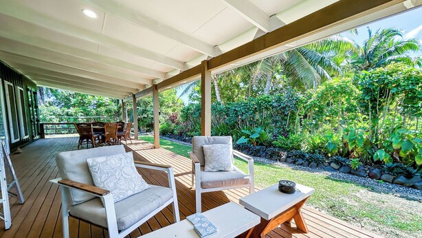 An outdoor covered patio with wooden flooring, white furniture, and a view of a lush garden. - Amori Villa & Cabin (Rarotonga)