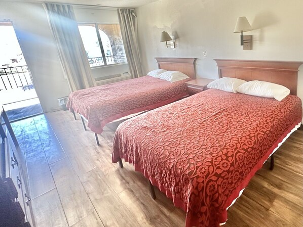 Habitación de hotel con dos camas, suelo de madera, balcón con barandilla y ventana con cortinas.