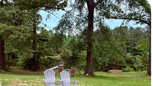 A fire pit with logs and two Adirondack chairs in a grassy outdoor area surrounded by trees. - 3-bedroom house w/pond next to White Oak Lake (Chidester)