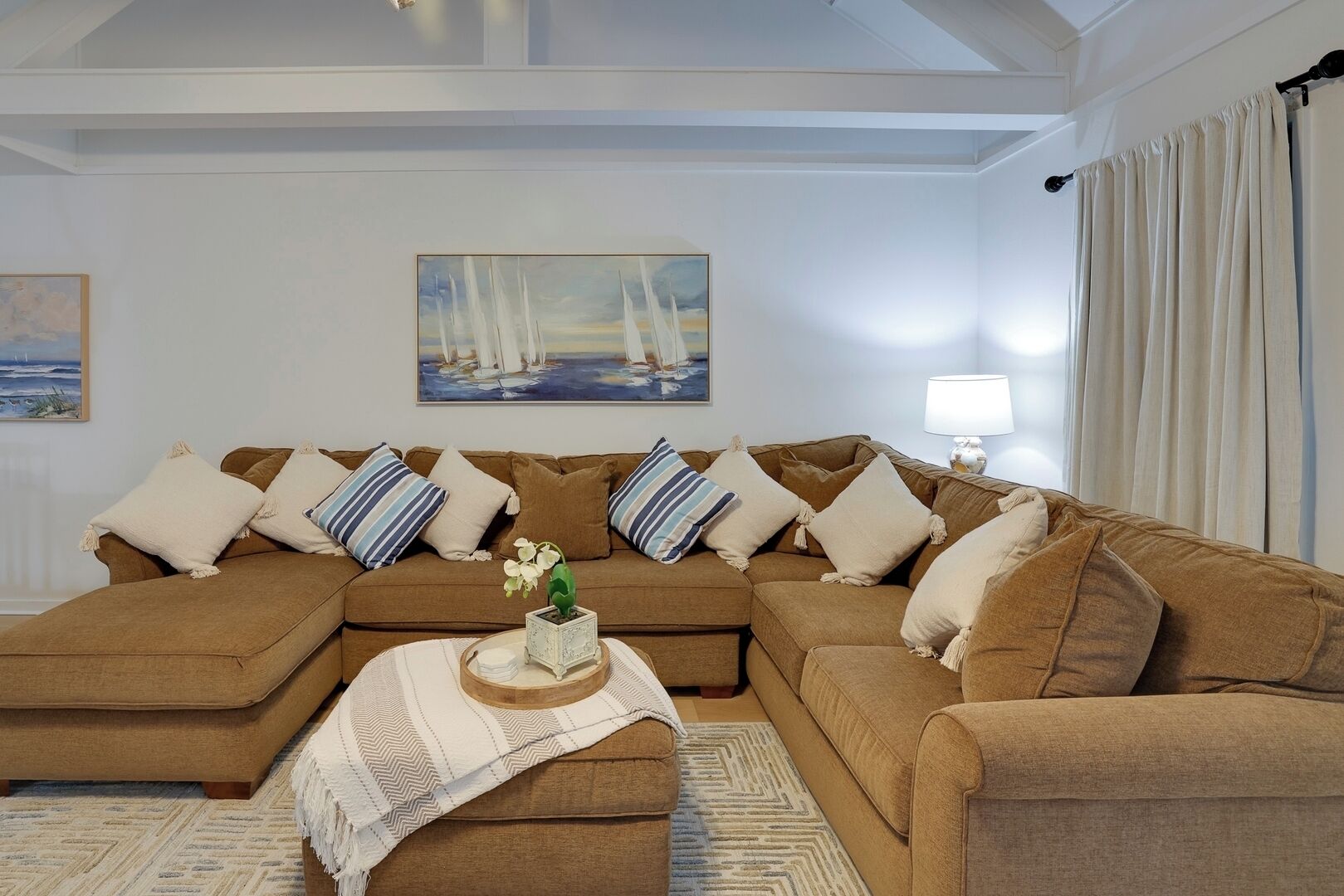 Un salón con un sofá seccional marrón, almohadas a rayas, una mesita con un jarrón y un cuadro de veleros en la pared.