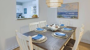 Un comedor con mesa de madera, sillas blancas, servilletas azules y un centro de mesa tejido.