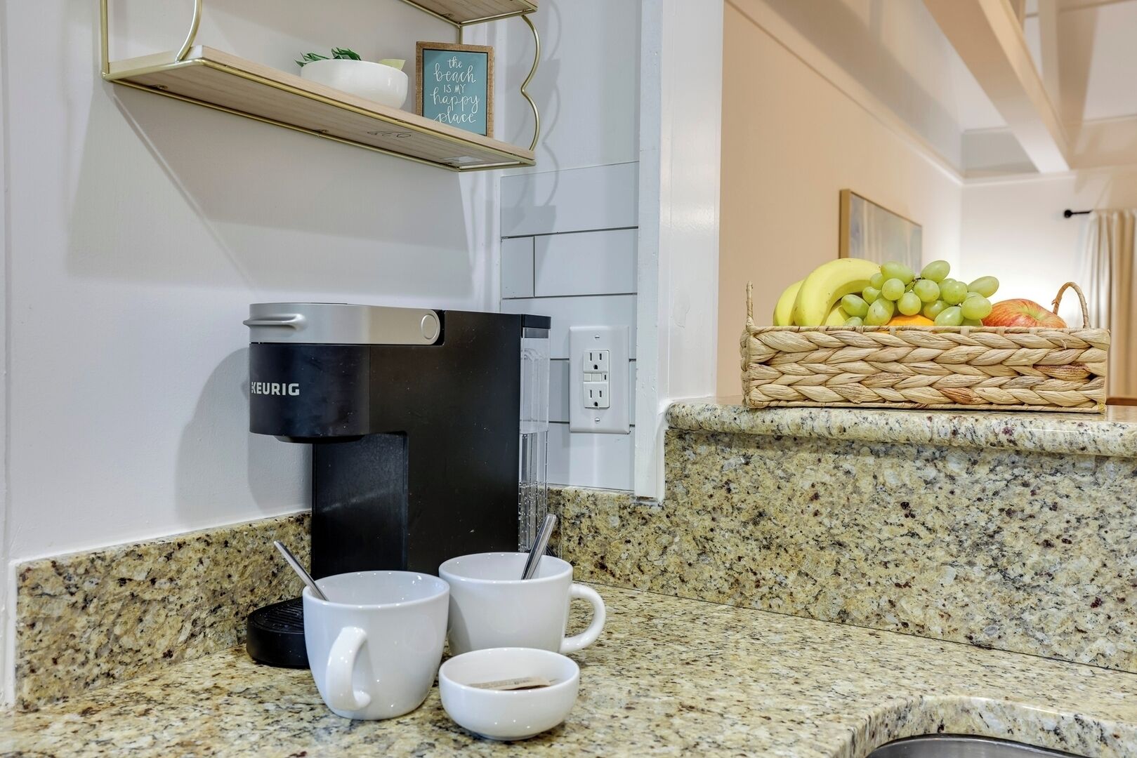 A kitchen with a coffee maker, two mugs, a basket of fruit, and a shelf with a decorative sign.