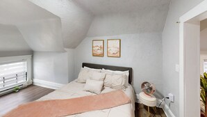 A bedroom with a sloped ceiling, a bed with multiple pillows, two wall-mounted paintings, a bedside fan, and a window with blinds.