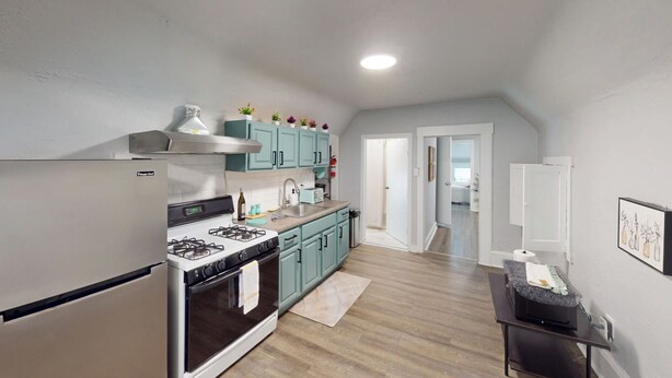 A modern kitchen with a gas stove, stainless steel appliances, teal cabinets, and a wooden floor.