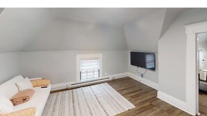 A room with a white ceiling, a flat-screen TV mounted on the wall, a white sofa with patterned cushions, and a patterned rug on the wooden floor.