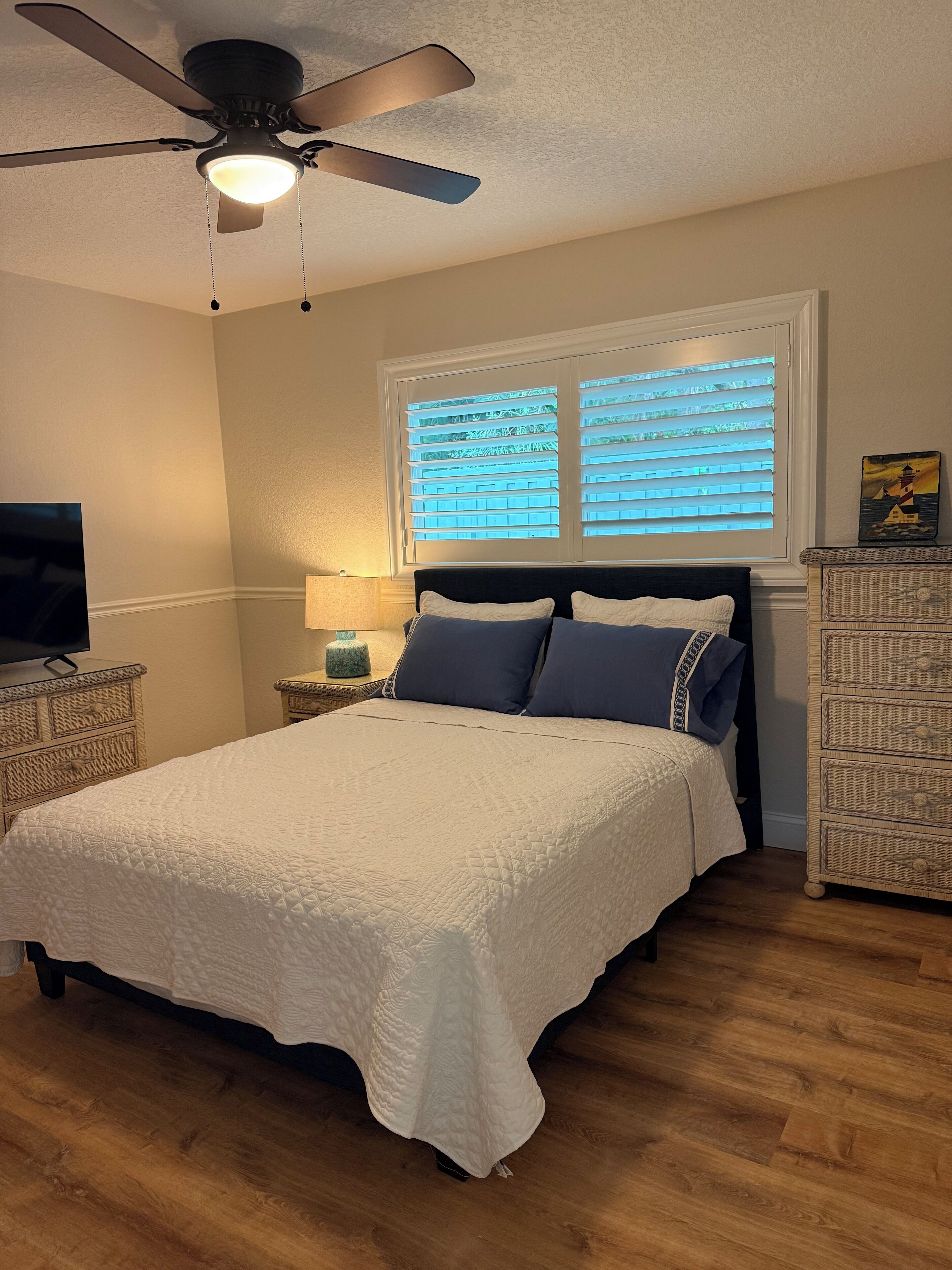 A bedroom with a bed, a ceiling fan, a window with blinds, a lamp, a dresser, and a television.