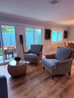 Una sala de estar acogedora con muebles de mimbre, una mesa de centro de vidrio y vistas a una valla azul en el exterior.