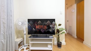 Una sala de estar con un televisor de pantalla plana sobre un soporte, un ventilador blanco, una planta en maceta y una puerta de madera al fondo.