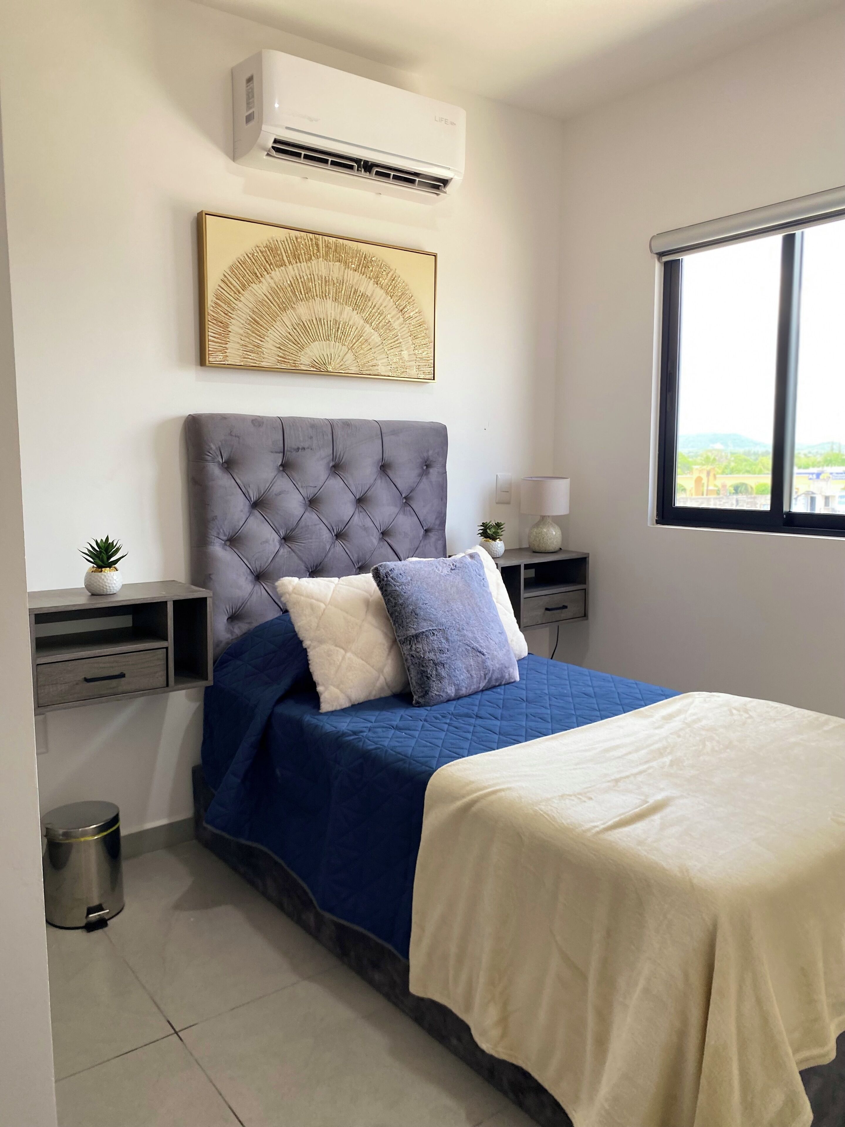 A bedroom with a large bed, a grey upholstered headboard, a wall-mounted air conditioner, a decorative wall piece, a bedside table with a lamp, and a window with a view.