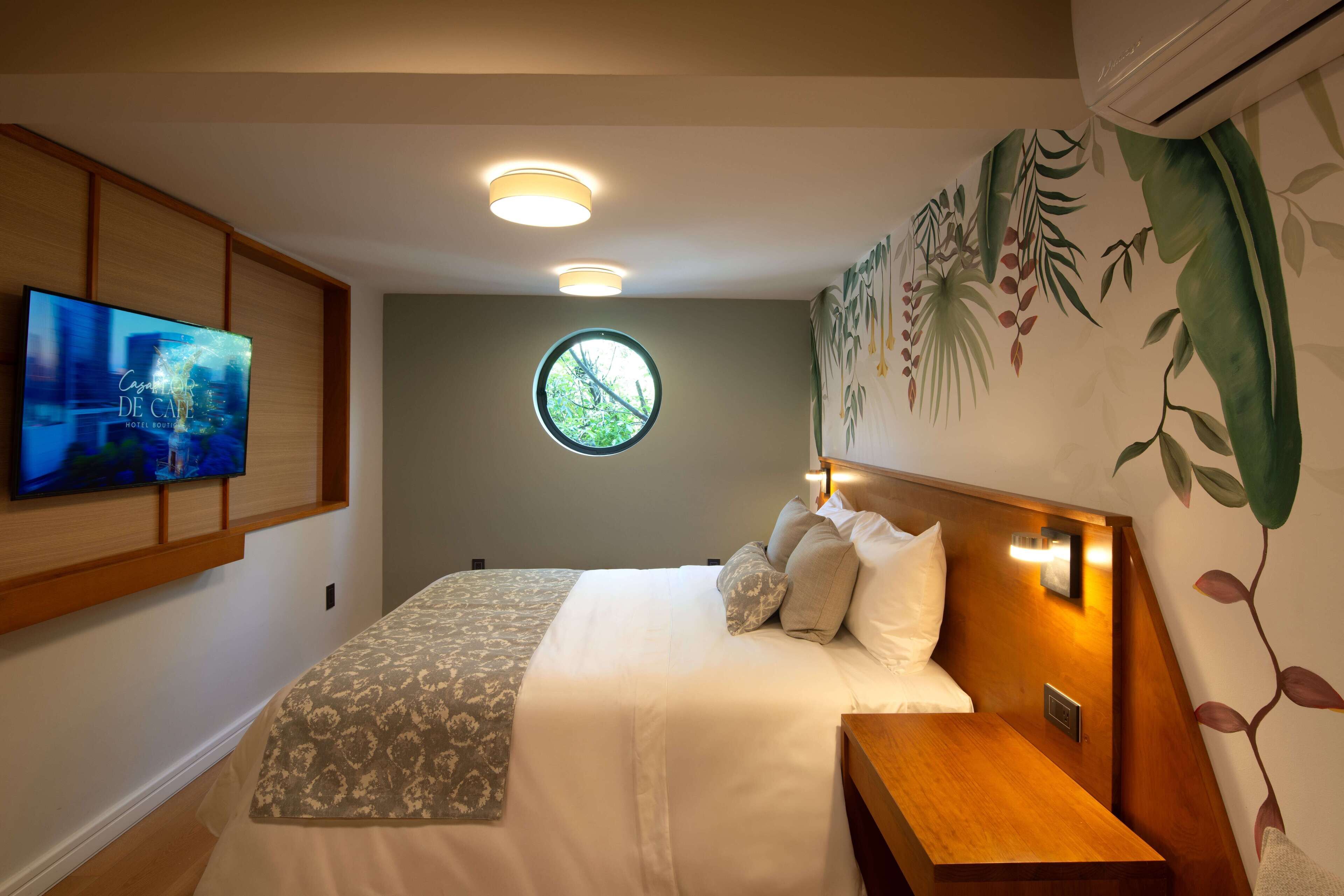 A modern hotel room with a large bed, a TV mounted on the wall, a round window, and a wall mural.